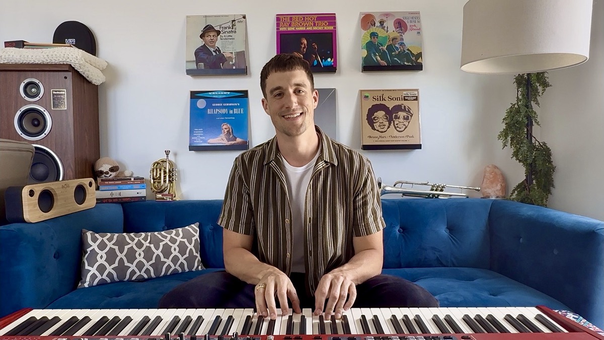 Greg smiling at a red Nord keyboard in his studio, seated on a blue velvet couch in front of a wall of jazz record sleeves.